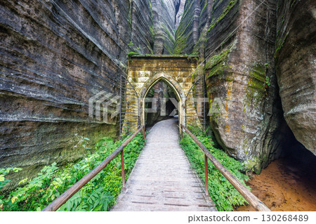 Adrspach-Teplice Rocks, sandstone formations in Hradec Kralove Region in Czech Republic, Europe. Pathway through a stone archway in a canyon landscape with lush greenery and towering rock formations. Adrspach-Teplice Rocks, sandstone formations in Hradec Kralove Region in Czech Republic, Europe. Pathway through a stone archway in a canyon landscape with lush greenery and towering rock formations. 130268489