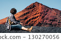 A girl sits on a black sand, enjoys a volcanic mountain landscape. 130270056