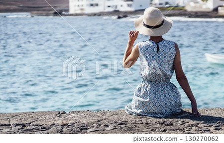 A girl in a blue dress with a hat on her head looks at the beautiful ocean on a sunny day. 130270062
