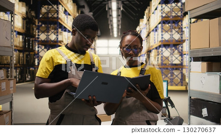 Black coworkers preparing orders in boxes for shipment in large warehouse, checking inventory on laptop and managing cargo distribution. Sorting packages for logistics management. Camera A. 130270637