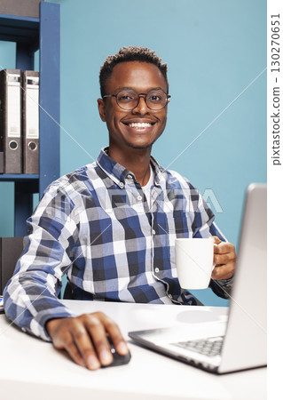 Portrait of smiling african american man with a coffee cup in hand, touching a mouse as he uses his laptop on desk. Black male entrepreneur holding a mug of hot drink and looking at camera. 130270651