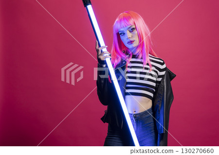 Caucasian female model stands in dramatic lighting, gripping vertical LED stick. Beautiful woman with pink hair and edgy striped outfit boldly posing with neon tube light against isolated background. 130270660