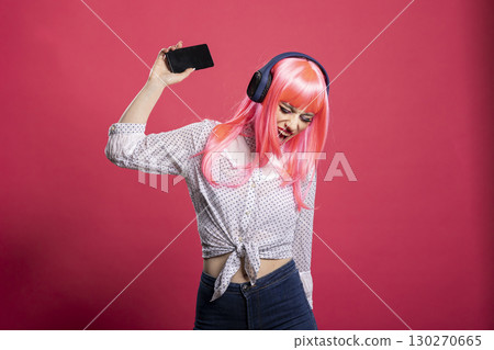 Energetic white woman with wireless headphones, joyfully singing and dancing with her arm in air, enjoying favorite music. Cheerful female music lover lost in the rhythm of her song playlists. 130270665