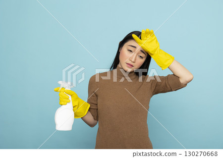Tired asian woman in yellow rubber gloves holding spray bottle, touching forehead in exhaustion after long cleaning chores. Housekeeping concept showing janitor with fatigue and stress. 130270668