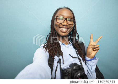 African american woman with DSLR around neck, showing peace sign to camera, filming content for vlogging channel. Cheerful black female photographer greets online viewers with two finger, v gesture. 130270713