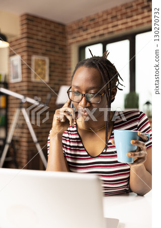 Focused entrepreneur works from home, reviewing product design on laptop while speaking to merchant on cellphone. Black woman holds coffee cup, engaged in business call during remote work session. Focused entrepreneur works from home, reviewing product design on laptop while speaking to merchant on cellphone. Black woman holds coffee cup, engaged in business call during remote work session. 130270732