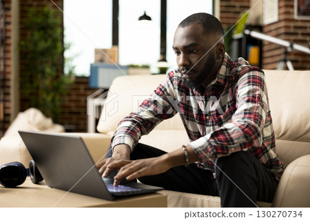 Serious male manager seated in cozy home office, typing on laptop and reviewing financial data. African american entrepreneur stays productive, working remotely on maintaining startup company growth. Serious male manager seated in cozy home office, typing on laptop and reviewing financial data. African american entrepreneur stays productive, working remotely on maintaining startup company growth. 130270734