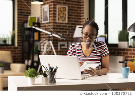 Young black woman prepares for project pitch, watching online course on laptop. Female freelancer seated at home office desk, writing in notebook, learning new skills through virtual training. 130270751