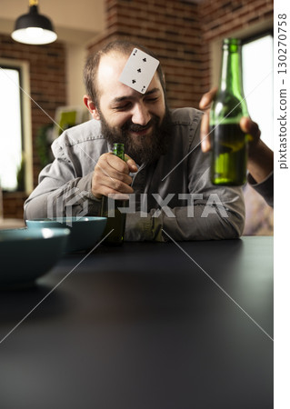Caucasian guy relaxes with friends, smiling with card on head and beer in hand during entertaining social gathering. Young white man enjoys bonding time, playing fun guessing game at table. 130270758