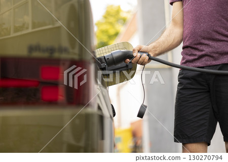 Close up of man charging electric vehicle at EVSE station, showcasing green energy transportation. Driver connecting EV plug to electric car to recharge it, concept of low emissions technology Close up of man charging electric vehicle at EVSE station, showcasing green energy transportation. Driver connecting EV plug to electric car to recharge it, concept of low emissions technology 130270794