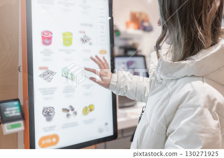 A woman is interacting with a digital menu displayed at a modern retail store today. A 30 -35-year-old woman in a white winter jacket orders food through the screen menu at a fast food restaurant 130271925