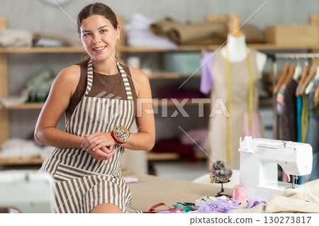 young dressmaker stands near the table and smiles young dressmaker stands near the table and smiles 130273817