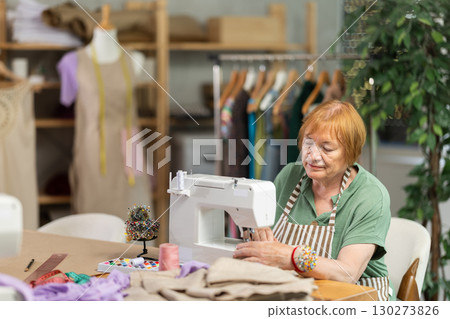 Mature female seamstress is sitting near sewing machine and create new dress in workshop Mature female seamstress is sitting near sewing machine and create new dress in workshop 130273826