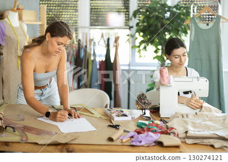 Female fashion designer sketching outfit while teammate sewing at machine 130274121