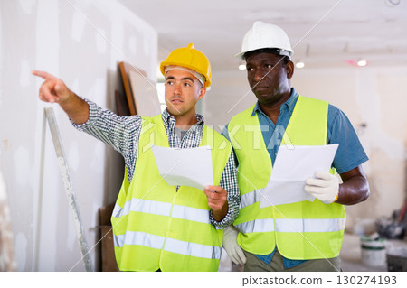 Foreman and worker in yellow vest and protective helmet are discussing the repair of a room in cottage 130274193