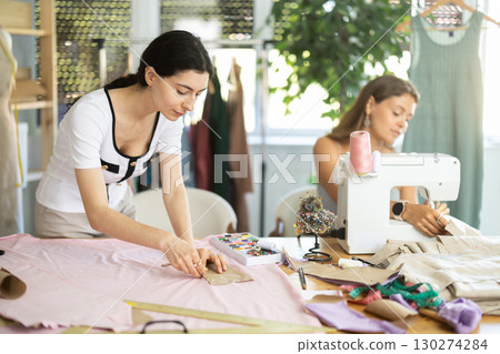 Female dressmaker cutting fabric next to assistant 130274284