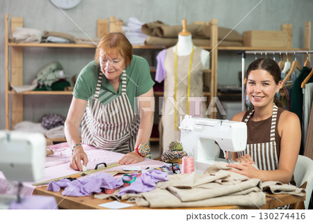 Elderly dressmaker making marks on fabric next to young seamstress Elderly dressmaker making marks on fabric next to young seamstress 130274416