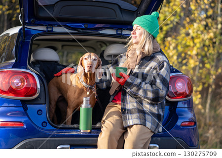 Woman pet owner drinking tea sitting in car trunk with dog enjoying nature in forest on weekend. 130275709