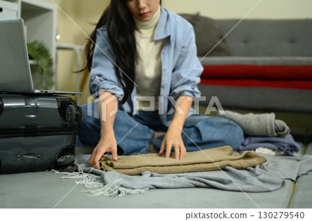 Young woman folding clothes on floor while packing suitcase for travel at home 130279540