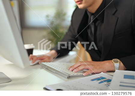 Businessman in black suit typing on keyboard while working with financial charts at desk 130279576