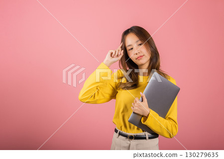 Portrait Asian young woman in a yellow shirt is smiling and holding up her laptop computer and thinking gesture studio shot isolated on pink background, Thai female think her work 130279635