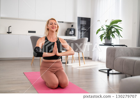 Middle aged blonde woman standing on yoga mat in namaste pose at home, focusing on breathing, mindfulness, and balanced posture 130279707