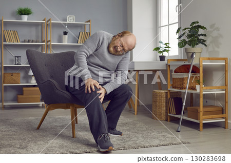 Senior man who suffers from knee pain is sitting on a chair and holding his knee 130283698