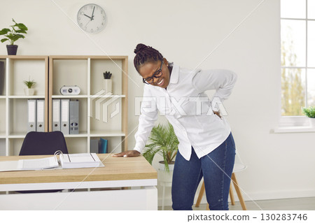 Woman standing by her working desk in office, feeling pain and holding hand on her lower back 130283746