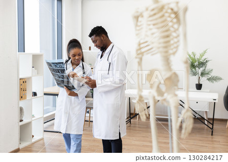 Medical professionals male and female discussing X-rays in modern clinic with skeleton model emphasizing anatomy and collaboration in medical settings Medical professionals male and female discussing X-rays in modern clinic with skeleton model emphasizing anatomy and collaboration in medical settings 130284217