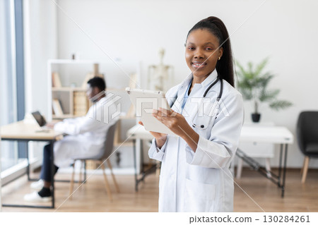 Young adult African female doctor holding tablet smiling confidently in medical clinic office. Male colleague working on computer in background. Bright modern health care workspace focused on teamwork Young adult African female doctor holding tablet smiling confidently in medical clinic office. Male colleague working on computer in background. Bright modern health care workspace focused on teamwork 130284261