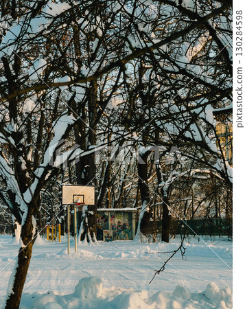 Snow covering basketball court in park during winter, with trees and graffiti wall in background, creating serene and desolate atmosphere. Snow blanketing empty basketball court, surrounding trees Snow covering basketball court in park during winter, with trees and graffiti wall in background, creating serene and desolate atmosphere. Snow blanketing empty basketball court, surrounding trees 130284598