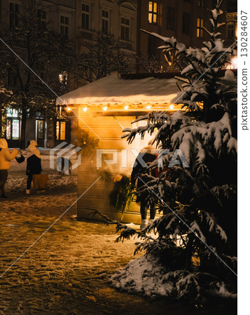 People are buying Christmas presents and decorations at illuminated wooden stall in snowy old town square at night, creating magical and festive atmosphere. Holiday shopper browse festive market stall 130284607