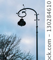Ornate street lamp adorned with icicles hangs gracefully on cloudy winter day, while bare tree branches create serene silhouette in foreground, enhancing tranquil atmosphere. Street lamp with icicles 130284610