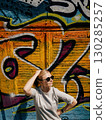 Woman with sunglasses and smartwatch posing with hand on hip and touching her hair against vibrant graffiti wall on sunny day, showcasing urban style and confidence. Woman and colorful graffiti wall 130285257