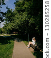 Young blond woman dressed in white clothing and cap, sitting on park bench, enjoying refreshing beverage on sunny summer day, surrounded by lush greenery and tranquility. Woman relaxing on park bench 130285258