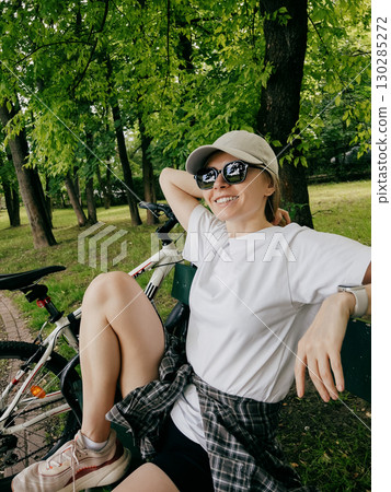 Young woman cyclist relaxing on park bench after an invigorating workout, soaking up fresh air and warm sunshine on beautiful summer day surrounded by nature. Female cyclist resting on park bench 130285272