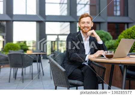 Smiling businessman with ginger hair sitting at urban cafe with laptop Smiling businessman with ginger hair sitting at urban cafe with laptop 130285577