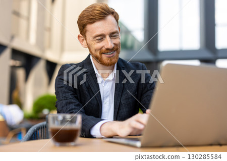 Smiling red-haired businessman in suit working on laptop at outdoor cafe 130285584