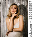 Portrait of young blonde woman wearing white dress and jewelry is posing touching her face in front of silver background during New Year Eve party. Female against shimmering silver party backdrop 130285897