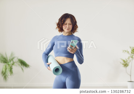 Sporty woman, yoga instructor standing holding mat checking mail, message on smartphone Sporty woman, yoga instructor standing holding mat checking mail, message on smartphone 130286732