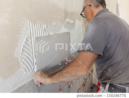 Craftsman places tile covered with adhesive on a bathroom wall, closeup. Construction or rennovation of a house Craftsman places tile covered with adhesive on a bathroom wall, closeup. Construction or rennovation of a house 130286778