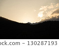 Golden sunlight peeks over mountain peaks at dusk, illuminating clouds and silhouetting trees in the valley 130287193