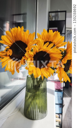 Bright yellow sunflowers in a ribbed green glass vase placed on a windowsill in a modern interior. The image conveys warmth, natural beauty and cheerful home decor 130288692