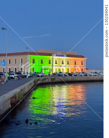 Coastal building illuminated in rainbow colors reflecting on the water during twilight, people casually sitting nearby. The vibrant lights create a striking visual effect Coastal building illuminated in rainbow colors reflecting on the water during twilight, people casually sitting nearby. The vibrant lights create a striking visual effect 130289041