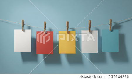 Colorful paper sheets hanging on string with wooden clothespins against blue wall, creating simple and organized display 130289286