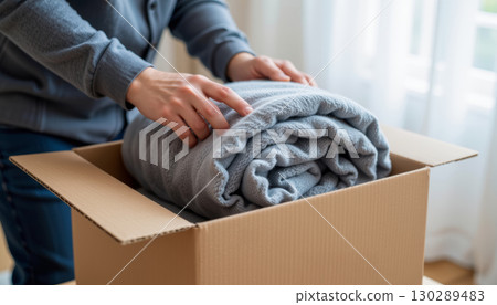 Person packing soft gray blanket into cardboard box, cozy home environment with natural light, careful hands folding fabric 130289483