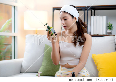 Beautiful young asian woman cheerful holding skincare or cream sitting on sofa at home. 130293021