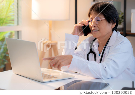 Senior asian doctor speak on smartphone while working on laptop in medical office. 130293038