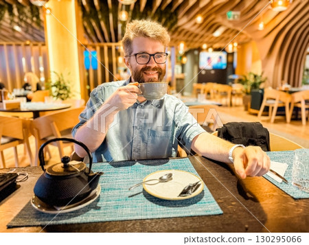 Man sipping hot tea at restaurant table with modern interior and warm lighting. Lifestyle, relaxation, leisure, and hospitality concept with cozy atmosphere and enjoyment. Man sipping hot tea at restaurant table with modern interior and warm lighting. Lifestyle, relaxation, leisure, and hospitality concept with cozy atmosphere and enjoyment. 130295066