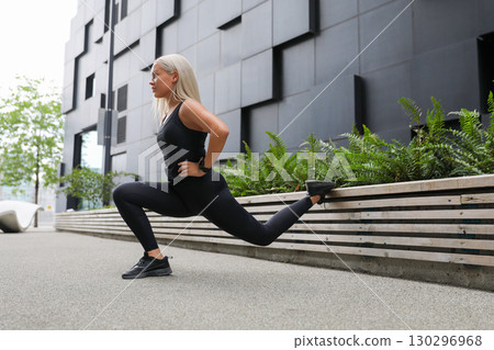 Active woman exercising outdoors with urban background, performing leg stretch lunge 130296968